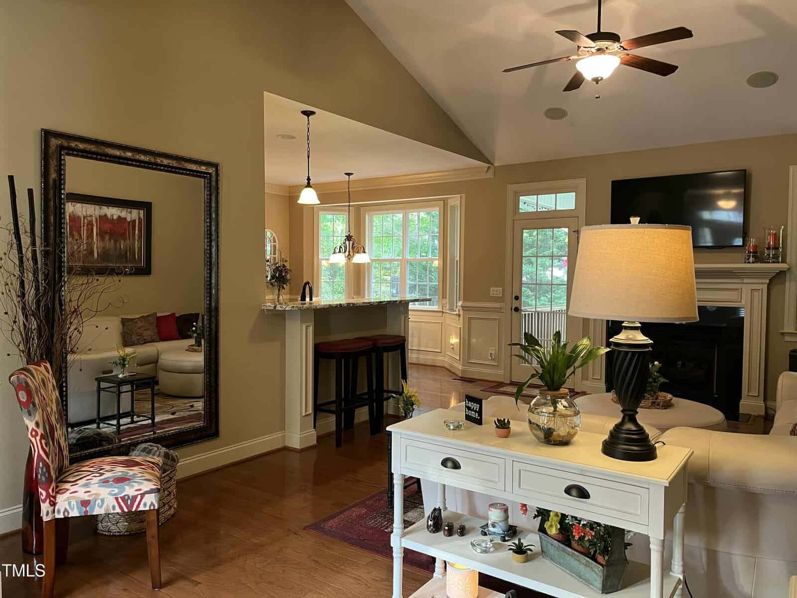 A living room with wood flooring, beige walls, and a vaulted ceiling. There is a white console table with a lamp, a mirror, a patterned chair, and a view into a kitchen with barstools and large windows.