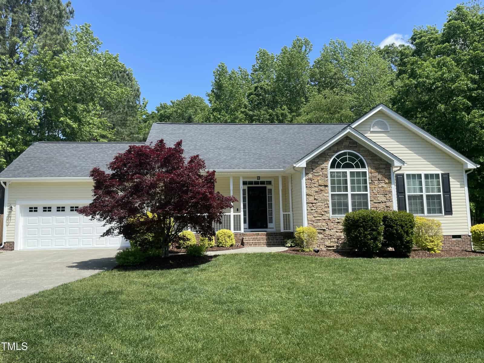 Single-story house with a two-car garage, light siding, stone accents, and arched window. There is a red-leafed tree, manicured lawn, bushes, and tall green trees in the background under a clear blue sky.