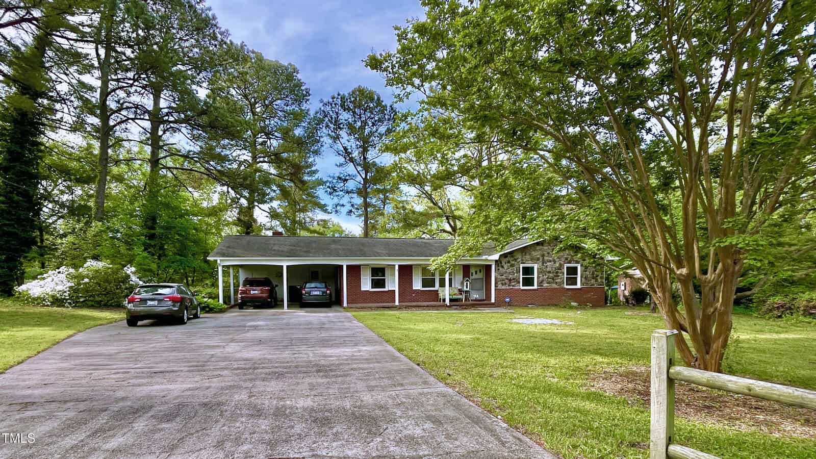 Single-story brick house with a covered carport, two cars parked, a large driveway, and surrounded by green grass, mature trees, and landscaping on a sunny day.