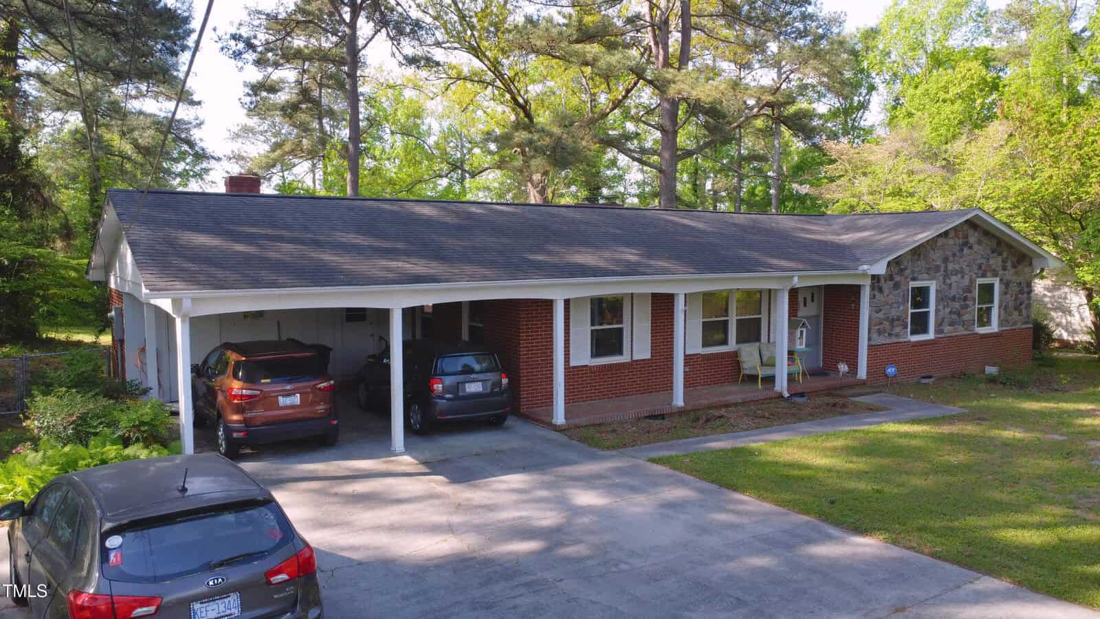 A single-story brick house with a dark roof, a stone facade section, and a two-car carport. Three cars are parked in the driveway and carport. Mature trees and greenery surround the house.