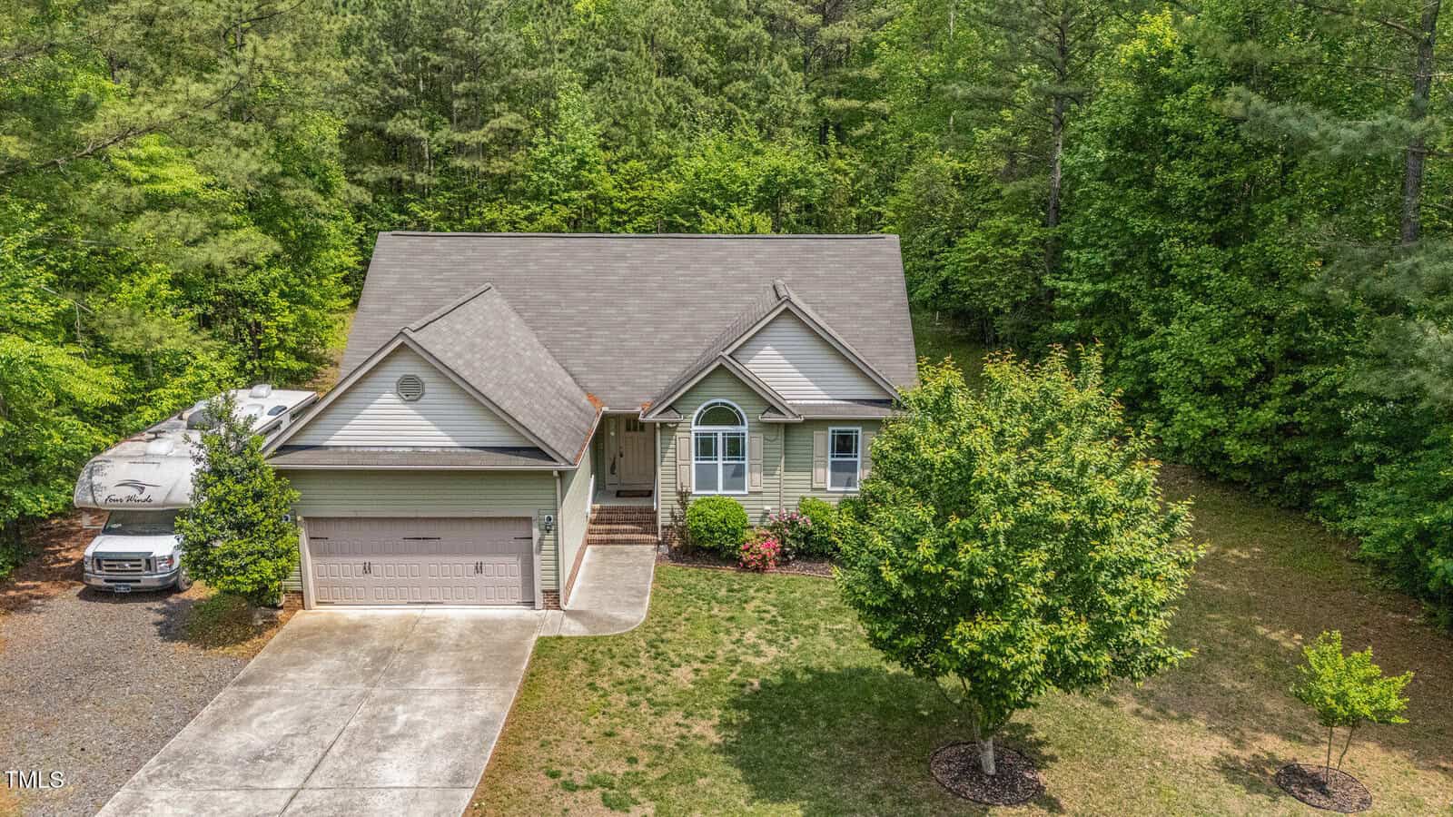 A light gray house with a two-car garage, pitched roof, and arched front window sits surrounded by green trees. A concrete driveway and walkway lead to the entrance, with a tree and bushes in the front yard.