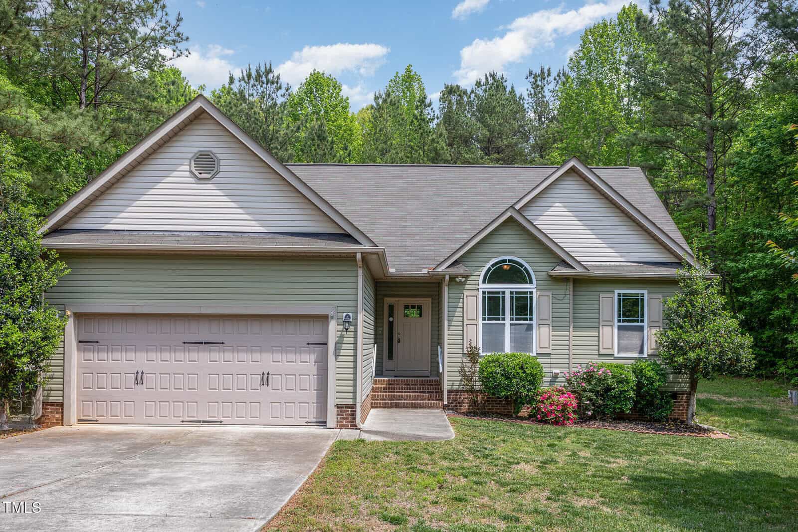 Single-story suburban house with a greenish exterior, gable roof, and two-car garage. The front yard has shrubs and flower beds, with a concrete driveway and a backdrop of tall trees under a blue sky.