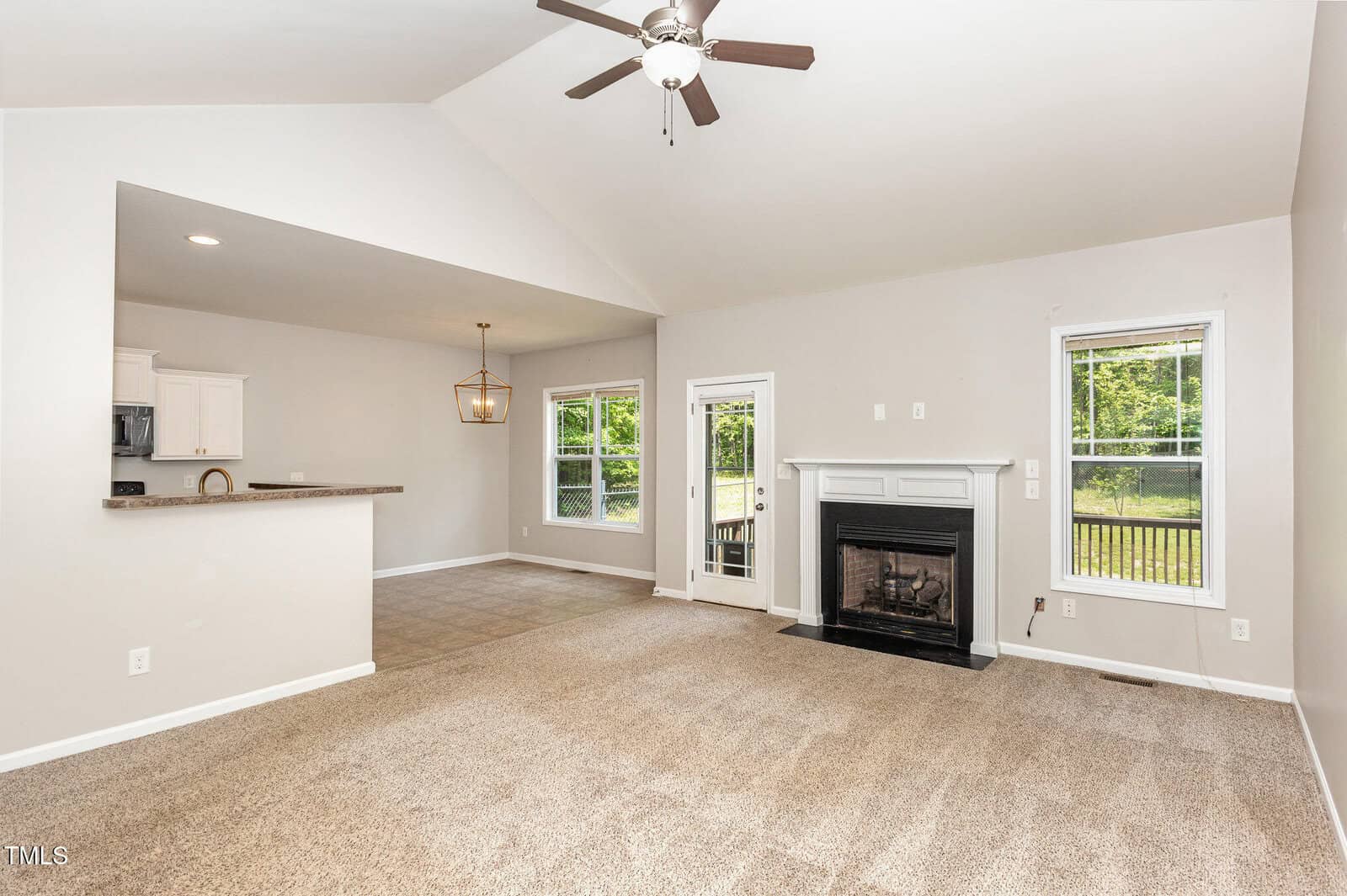 Bright, open living area with beige carpet, a ceiling fan, white walls, black fireplace, and large windows. The room connects to a dining area and kitchen with white cabinets and a breakfast bar.