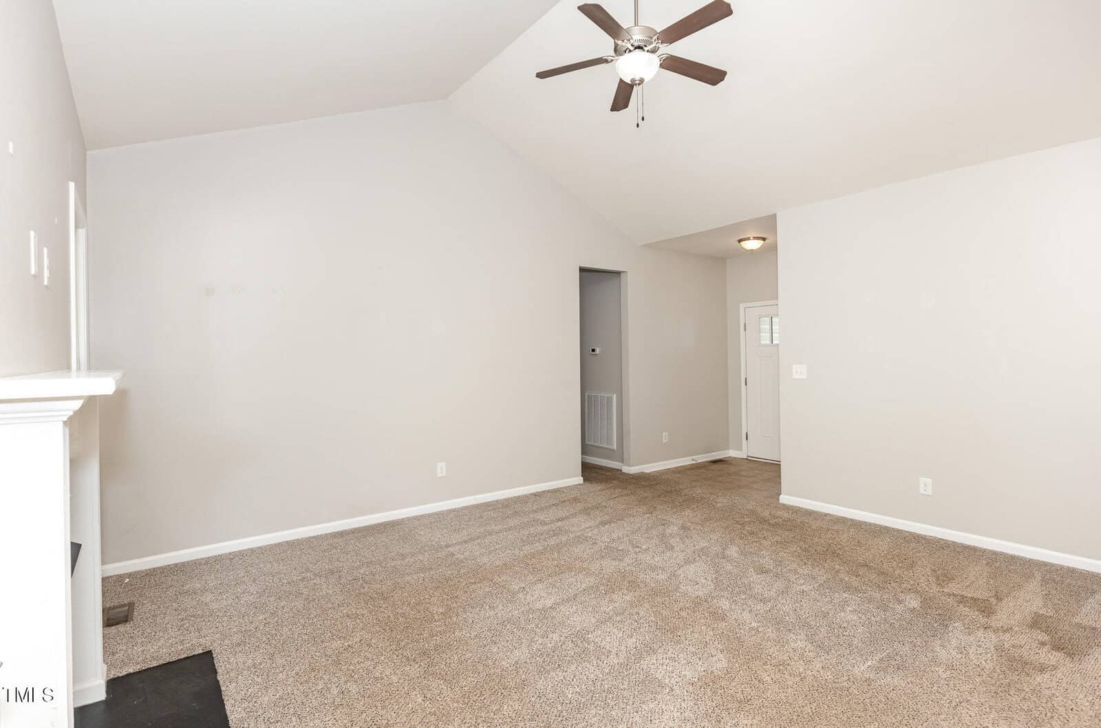 A bright, empty living room with beige carpet, light gray walls, a white fireplace on the left, a ceiling fan with lights, and a view of the entryway and front door in the background.