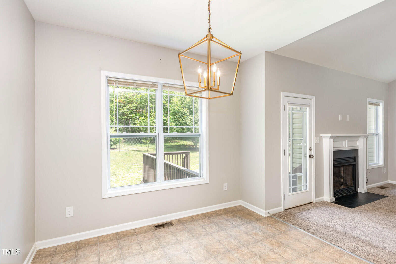 A room with a tile floor, large window, gold geometric light fixture, beige walls, and a carpeted area with a fireplace and door leading outside. Some greenery and a wooden deck are visible through the window.