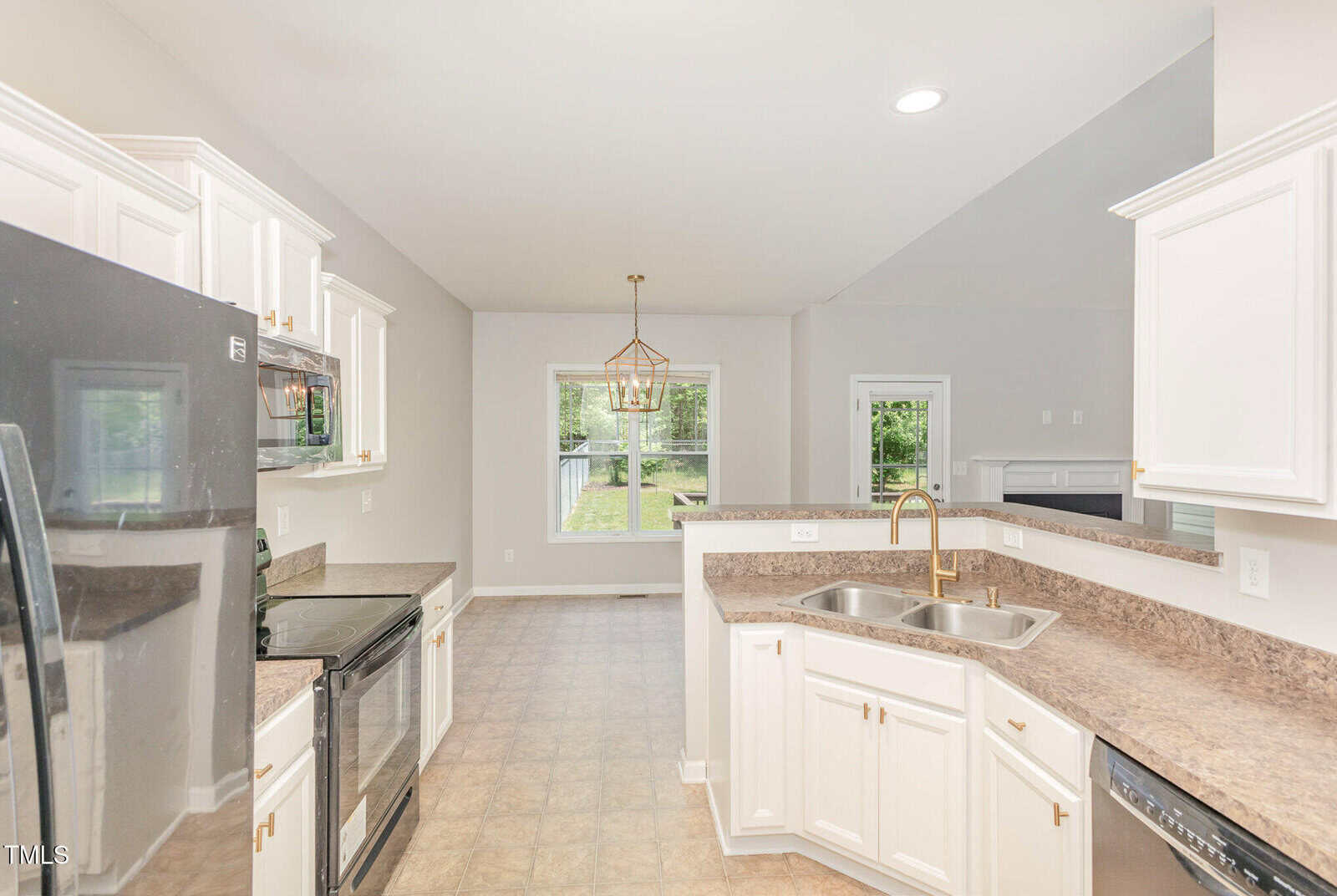 Modern kitchen with white cabinets, black appliances, double sink, and brown countertops. Open layout leads to a dining area with a window, gold chandelier, and a door to the outside. Neutral walls and tile flooring throughout.