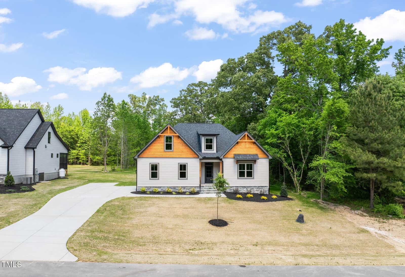A modern single-story house with gray siding and wood accents on the gables, a neatly landscaped yard, a curved driveway, and surrounded by lush green trees under a partly cloudy sky.