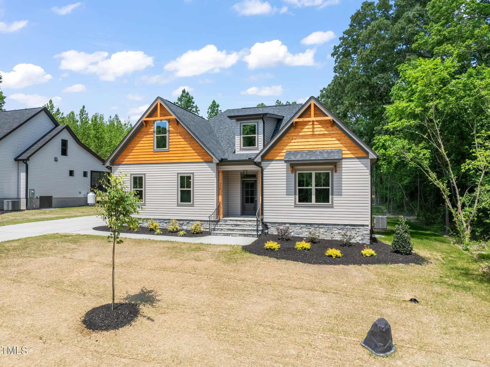 Modern house with light gray siding, dark gray roof, stone accents, and wood panel details. Small tree and fresh landscaping in the front yard, surrounded by grass and bordered by trees. Bright, sunny day with blue sky and clouds.
