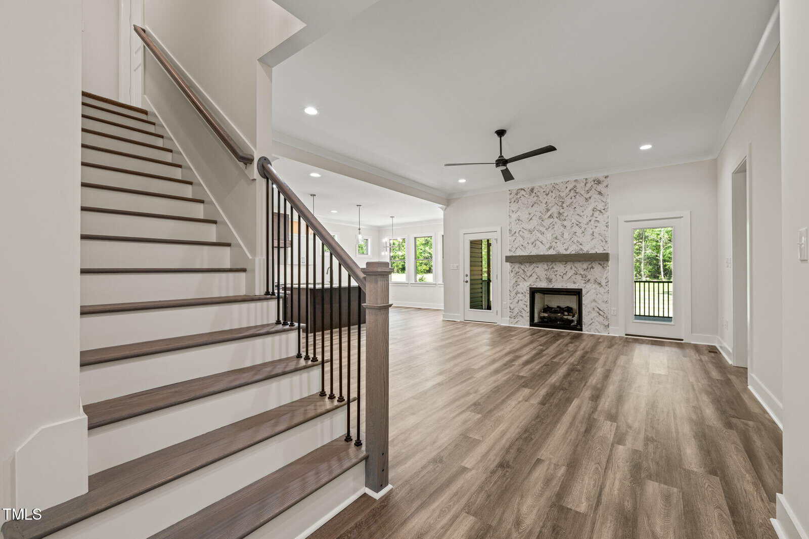Modern open-concept living room with wood flooring, a staircase with dark railing, ceiling fan, fireplace with patterned tile surround, large windows, and glass doors leading to a porch. Bright and spacious interior.