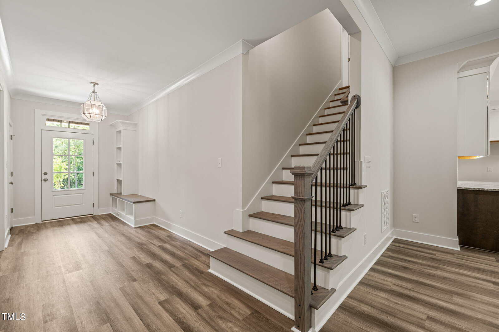 A modern entryway with wood flooring, white walls, a staircase with wooden steps and black balusters, built-in shelves and bench, and a glass front door allowing natural light inside.