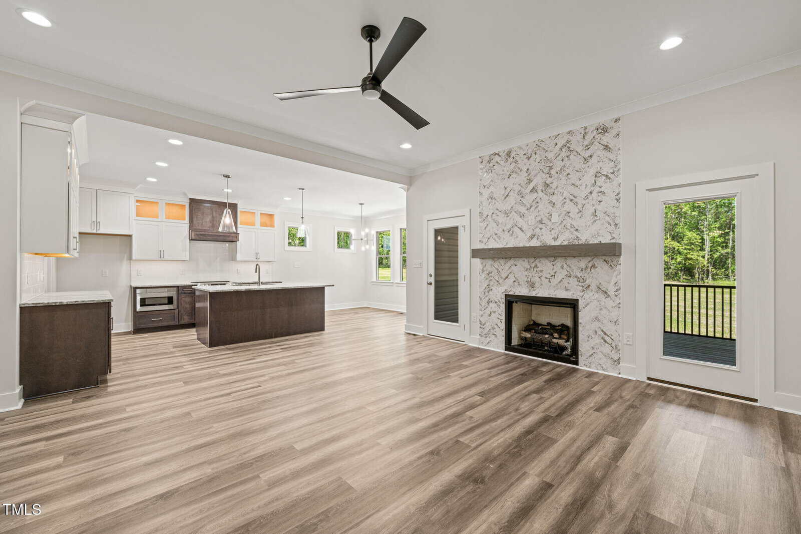Open-concept living area with wood floors, modern kitchen with island, white cabinetry, and stainless appliances. Fireplace with patterned tile surround, ceiling fan, doors leading outside, and large windows letting in natural light.