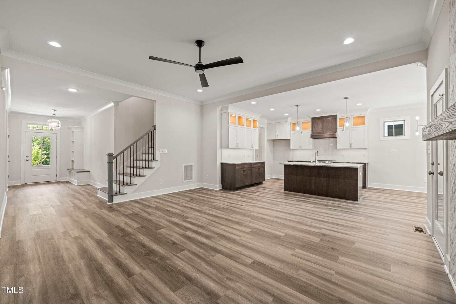 Open-concept living space with light wood flooring, white walls, modern ceiling fan, kitchen island with dark cabinets, stairway to the left, and front door visible in the background.