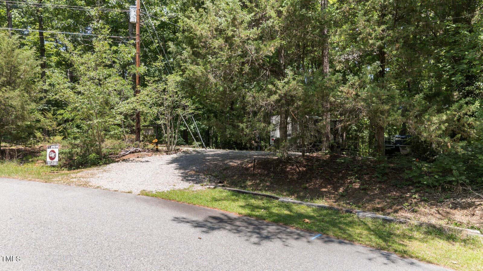 A gravel driveway leads into a wooded area with dense trees and greenery. A small sign near the road reads "posted" in red letters. Power lines and a utility pole are visible along the road.