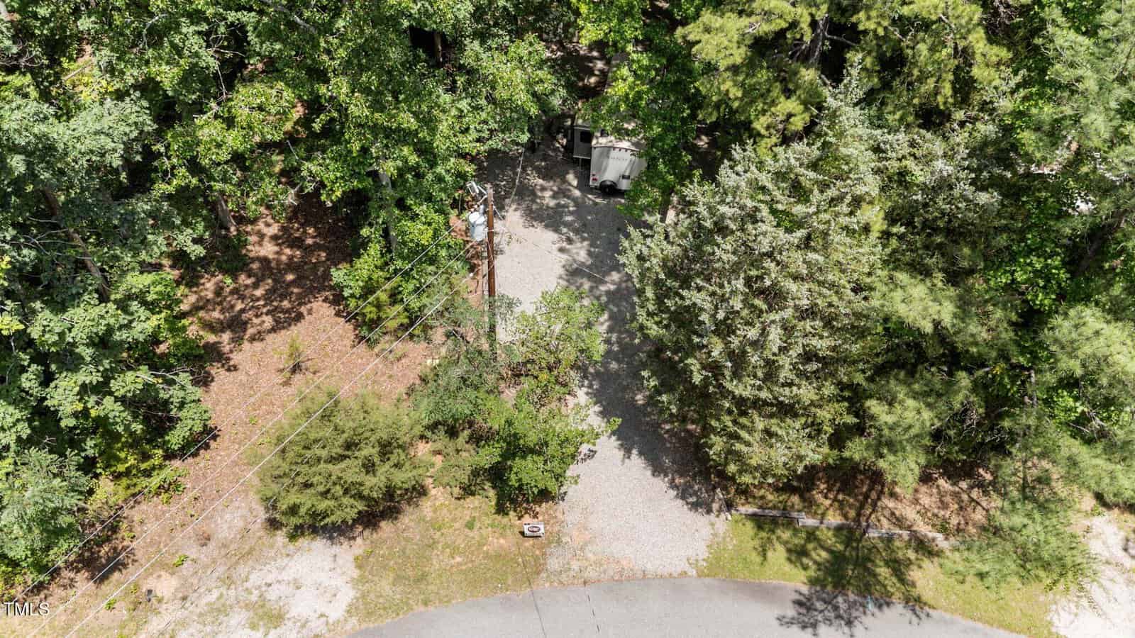 Aerial view of a gravel driveway surrounded by dense green trees, leading to a small shed or structure partially hidden by foliage near the end of the driveway, with a curved road visible at the bottom.