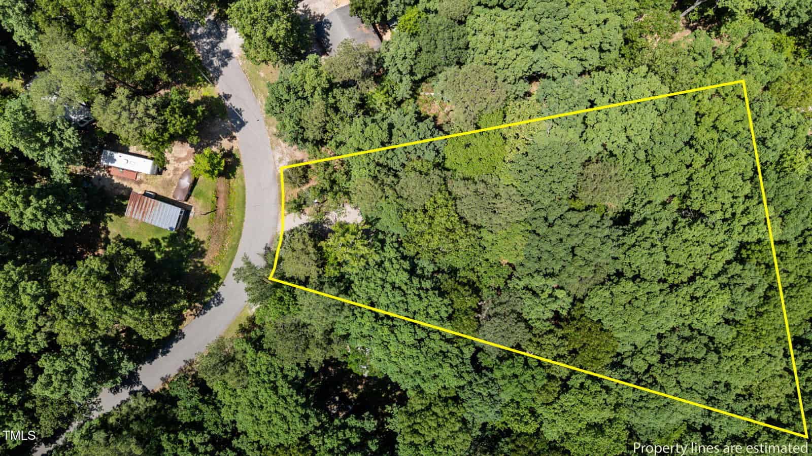 Aerial view of a wooded lot with estimated property lines outlined in yellow, adjacent to a curved road. A small building and a parked vehicle are visible opposite the lot, surrounded by dense trees.