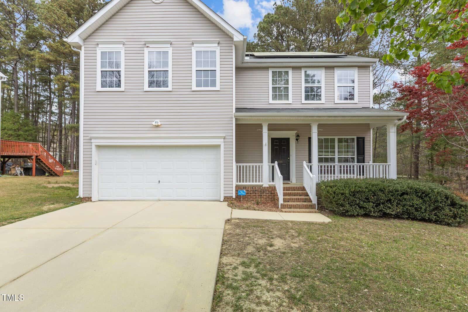Two-story beige house with white trim, a double garage, a covered front porch with railings, and a driveway. The yard is grassy with some trees and a deck visible in the background on the left.