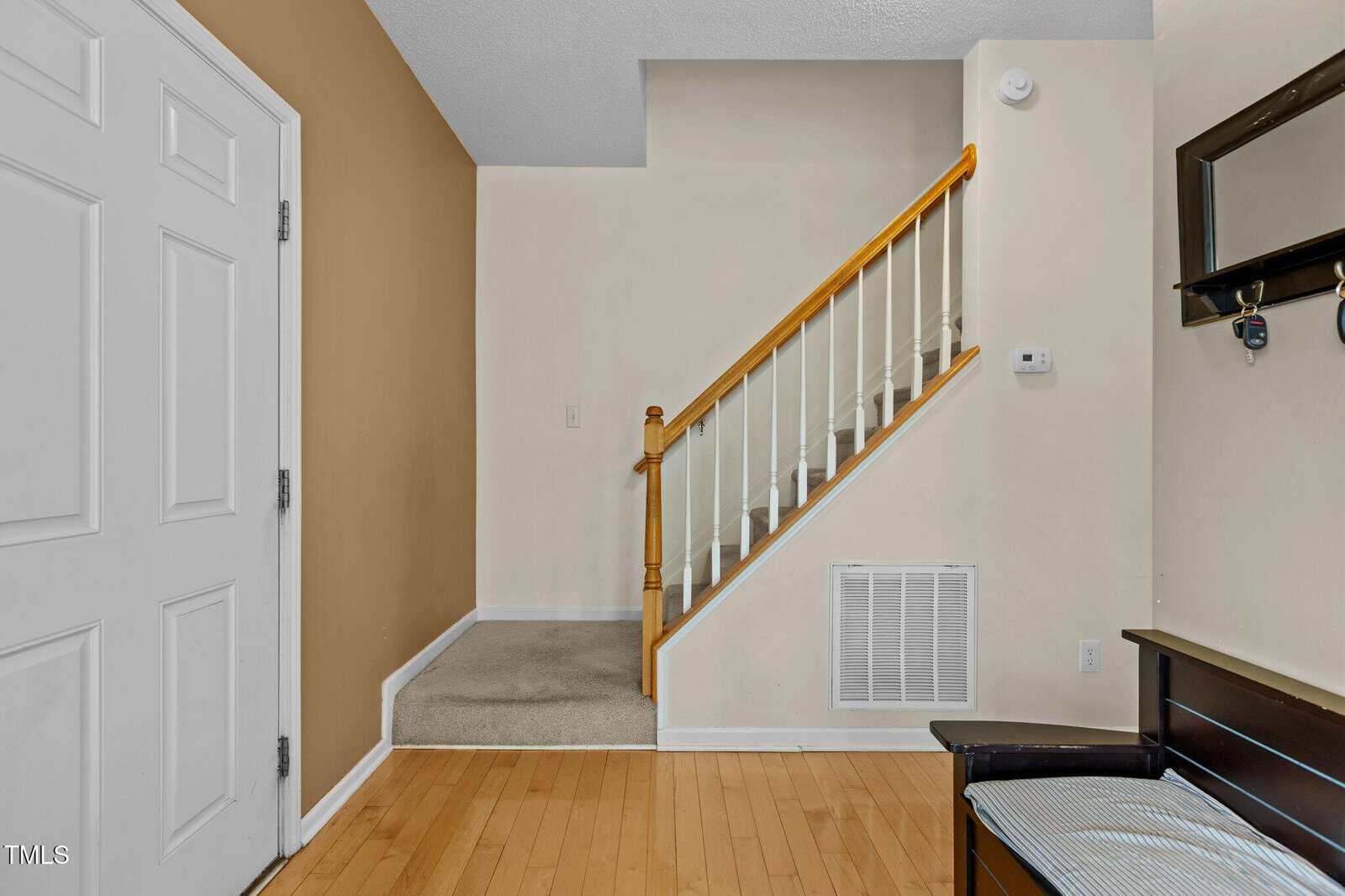 A view of a home entryway featuring a white front door, hardwood floor, a staircase with a wooden handrail, beige and white walls, a vent, and a black bench with a cushion and wall shelf nearby.