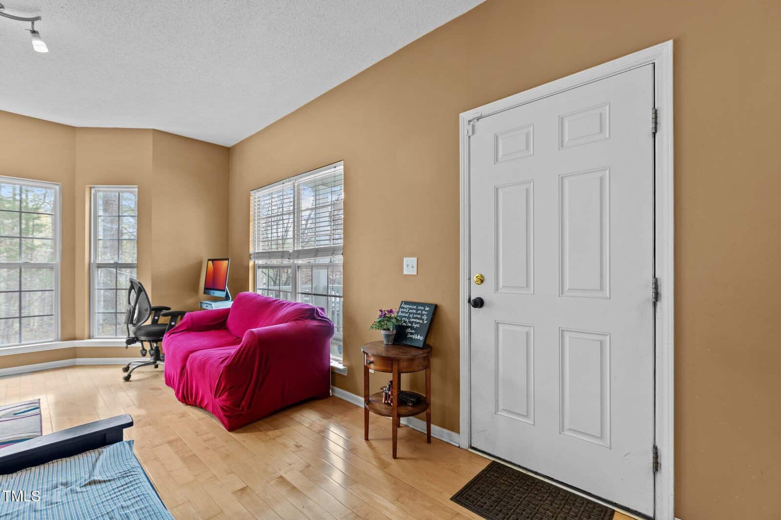 A room with tan walls and light wood floors features a white front door, a pink armchair, a desk with a computer, large windows, and a small table with flowers and a chalkboard.