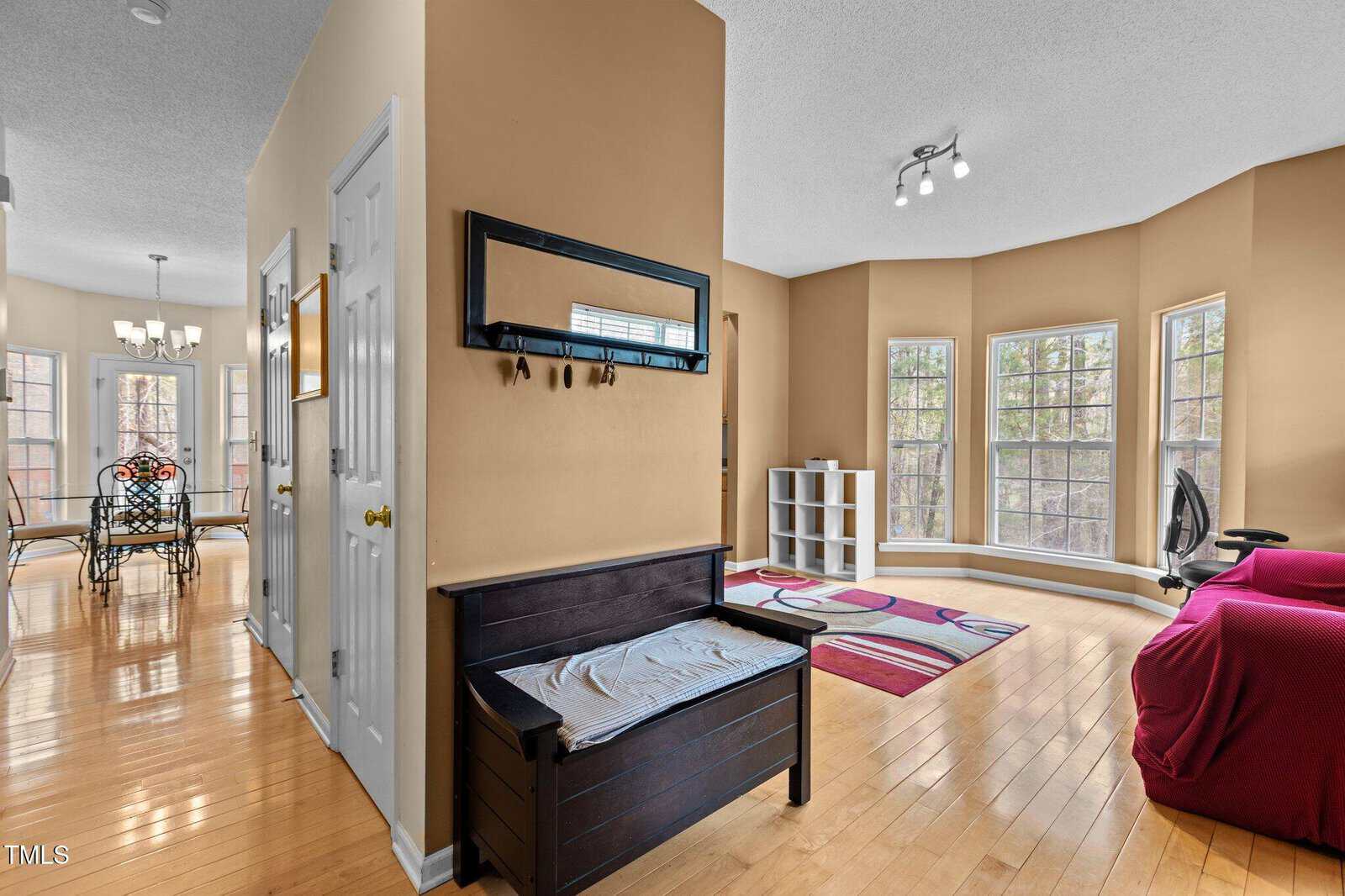 A hallway with tan walls leads to a dining area with a glass table and chairs. The entryway has a black bench with a mirror above it. The living space features large windows, a red sofa, and a colorful rug on hardwood floors.