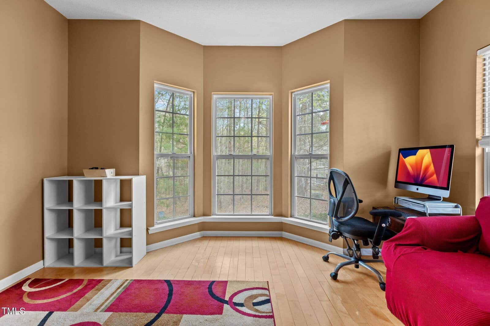 A home office with a bay window, a white cubed shelf, a black office chair, a desk with an iMac, a red sofa, and a patterned rug on a light wood floor. Trees are visible outside the windows.