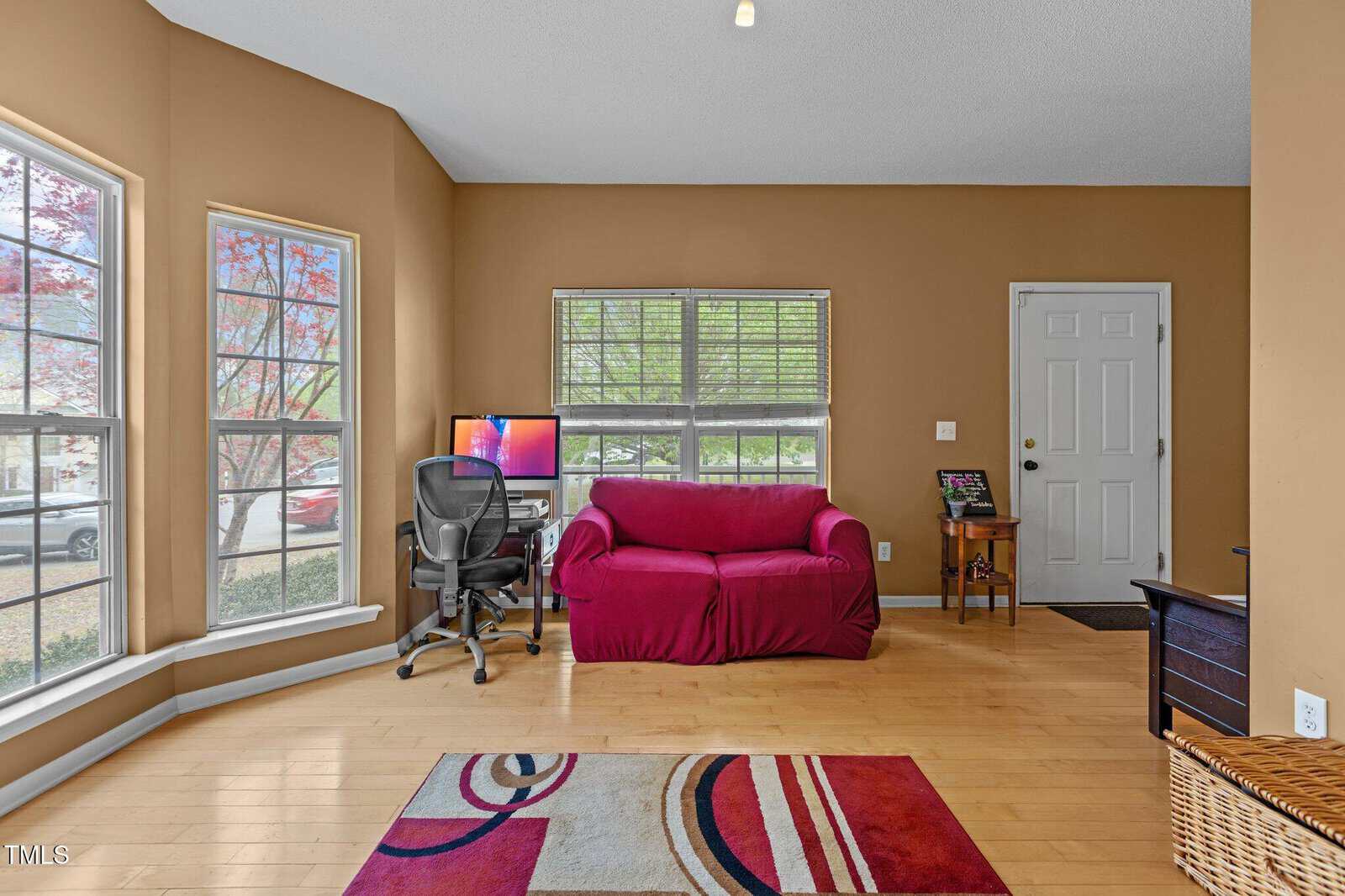 A living room with tan walls, light wood floors, a red loveseat, a modern desk with a computer and office chair by three tall windows, a small wooden side table, and a multicolored rug.