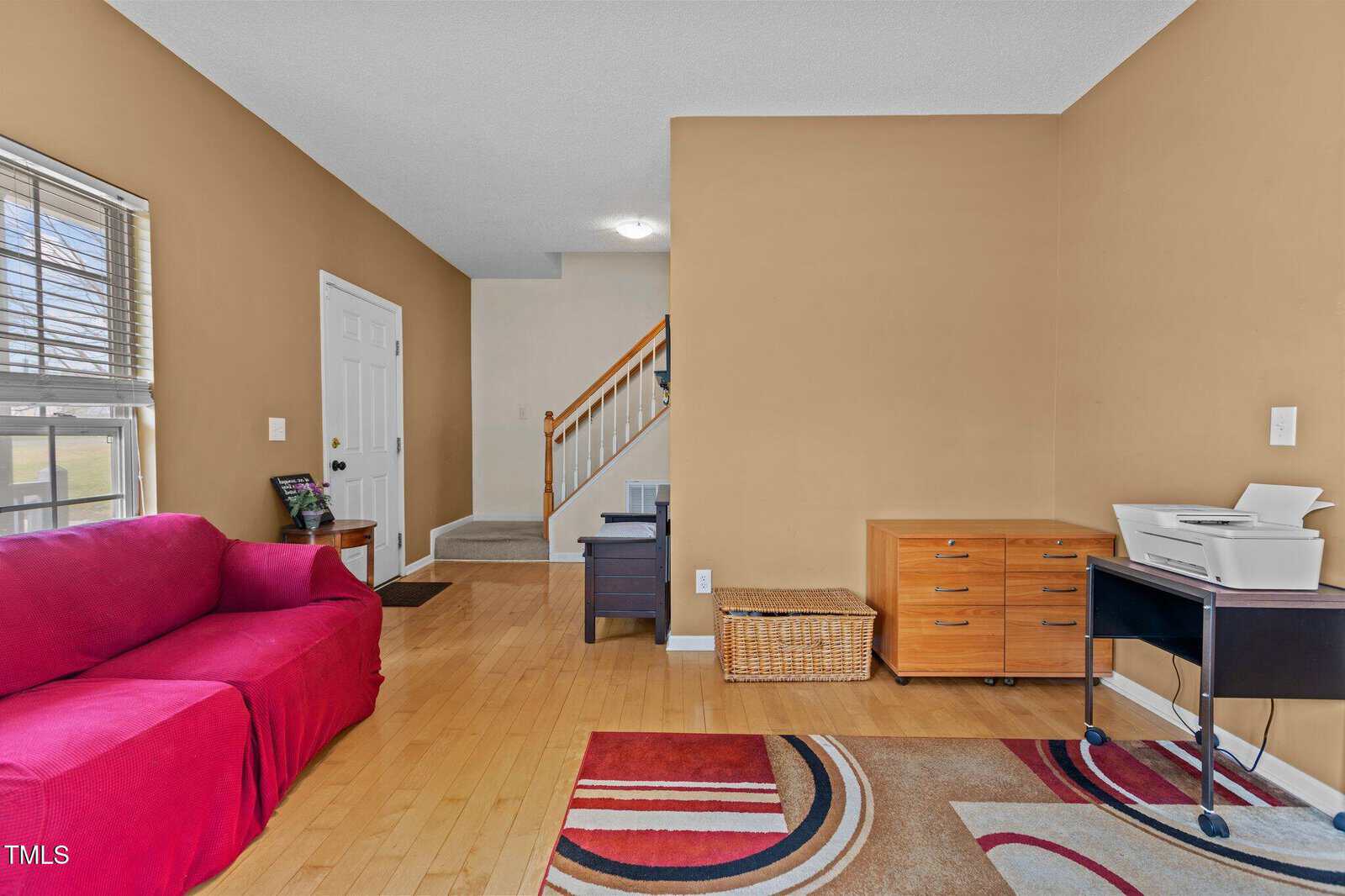 A living room with tan walls, wood flooring, a red sofa, a patterned rug, a wooden chest, a basket, a filing cabinet, a printer on a cart, and a staircase leading to the upper floor near the front door.