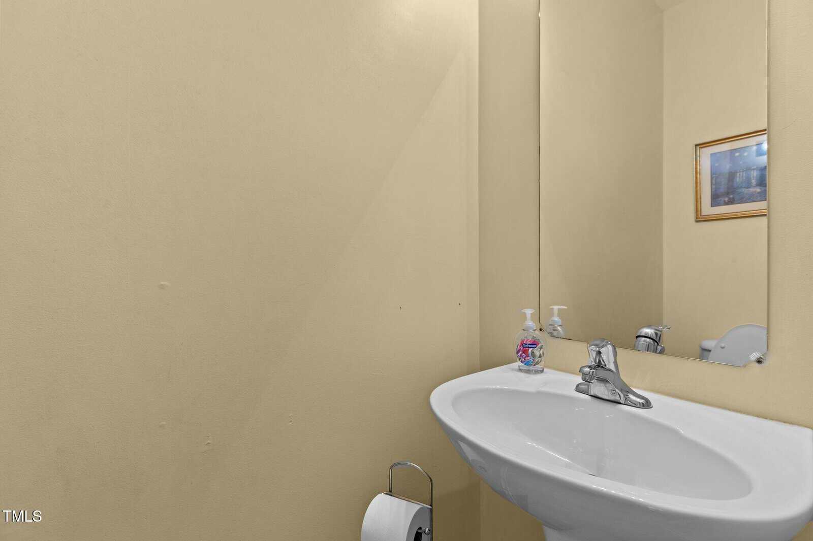 A small bathroom with beige walls, a wall-mounted oval mirror, a white sink with a silver faucet, a soap dispenser, a toilet partially visible, and a framed picture hanging on the wall.
