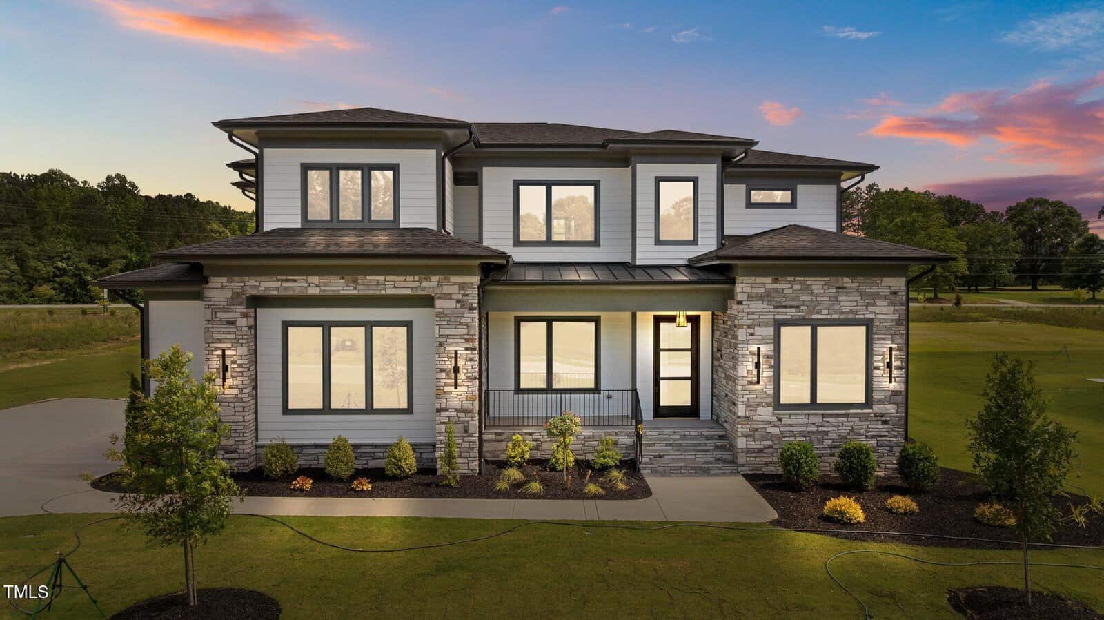 Modern two-story house at 200 Broadleaf Lane, Youngsville, with large windows, stone and siding exterior illuminated at sunset; neatly landscaped front yard features small shrubs and a green lawn.
