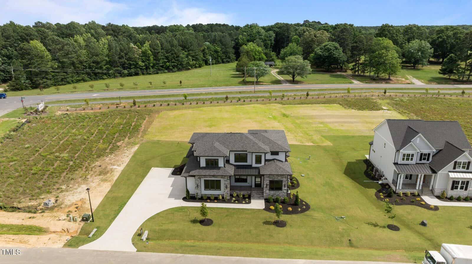 A large modern house with a gray roof and neatly landscaped yard sits on a spacious lot, with another house nearby and open grassy fields, trees, and a road in the background.