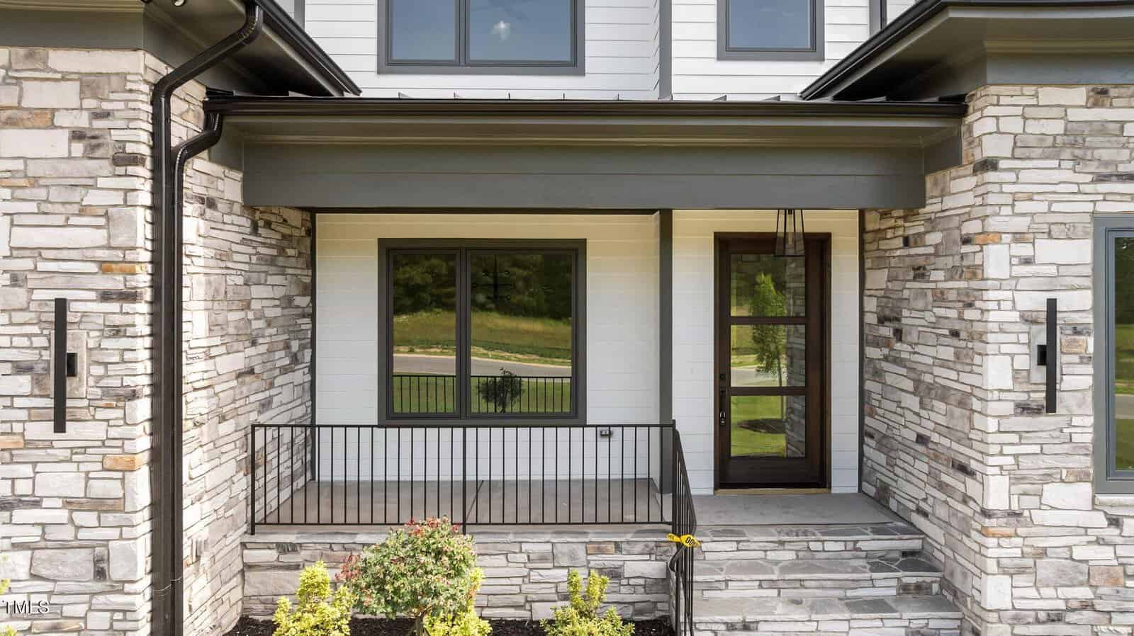 Modern house exterior featuring stone and white siding, a small covered front porch with black metal railing, large windows, a glass door, and landscaped bushes in front.