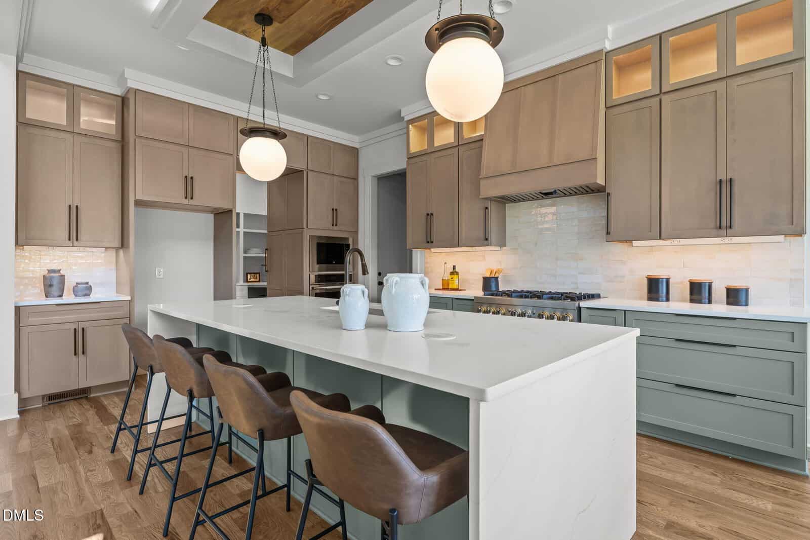 Modern kitchen with a large white island, four brown leather bar stools, pendant lights, light wood cabinets, stainless steel appliances, and two decorative vases on the island. Light wood floors and neutral tones throughout.