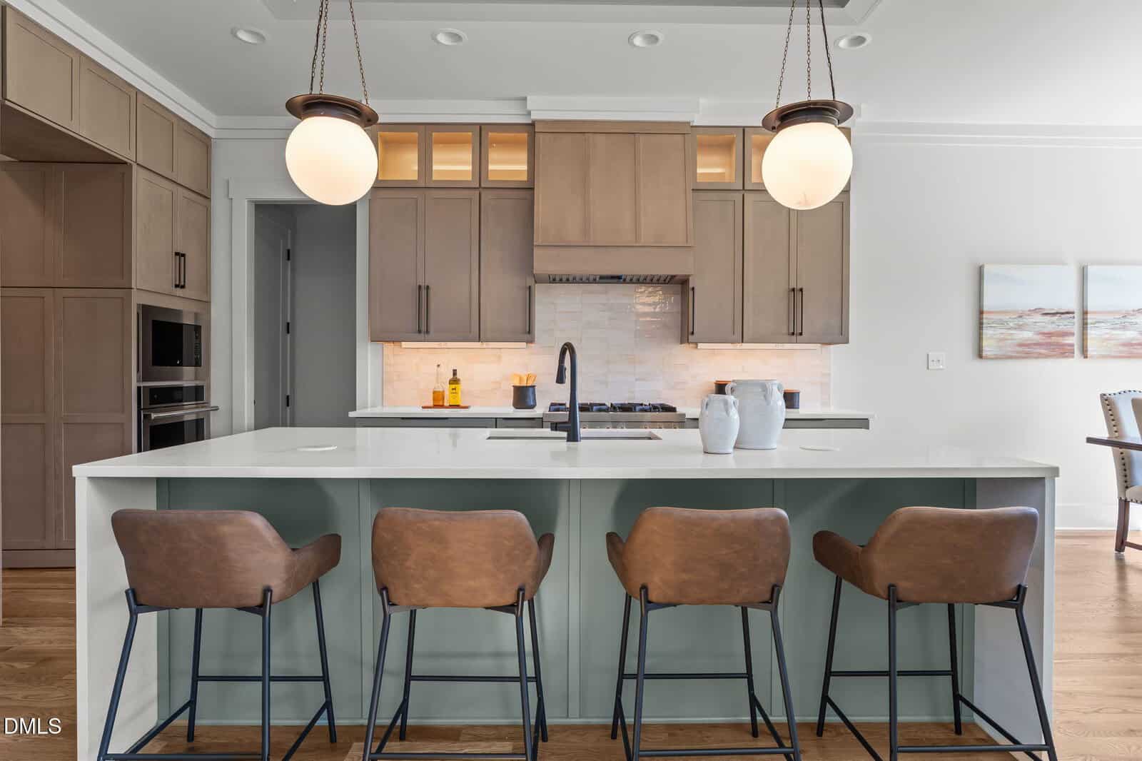 Modern kitchen with a large island, four brown leather barstools, light wood cabinets, built-in appliances, white countertops, two round pendant lights, and artwork on a wall in the background.