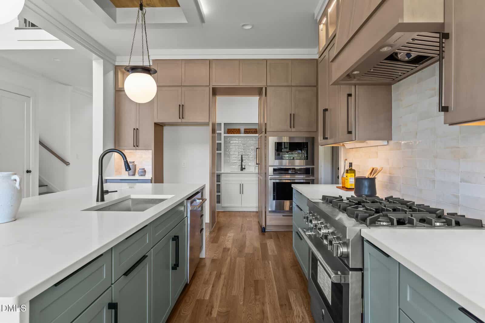 Modern kitchen with light wood floors, green lower cabinets, and white countertops. Stainless steel appliances, including a gas stove and double ovens, are built into the cabinetry. Subway tile backsplash and pendant lighting overhead.