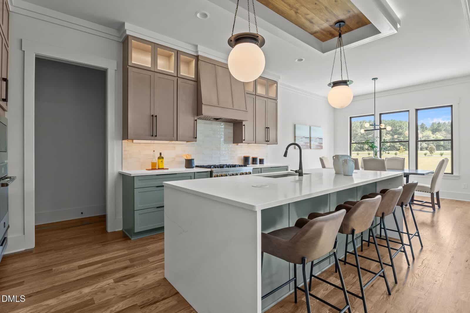Modern kitchen with a large white island, four cushioned barstools, pendant lights, wood floors, brown and sage green cabinetry, and large windows letting in natural light.