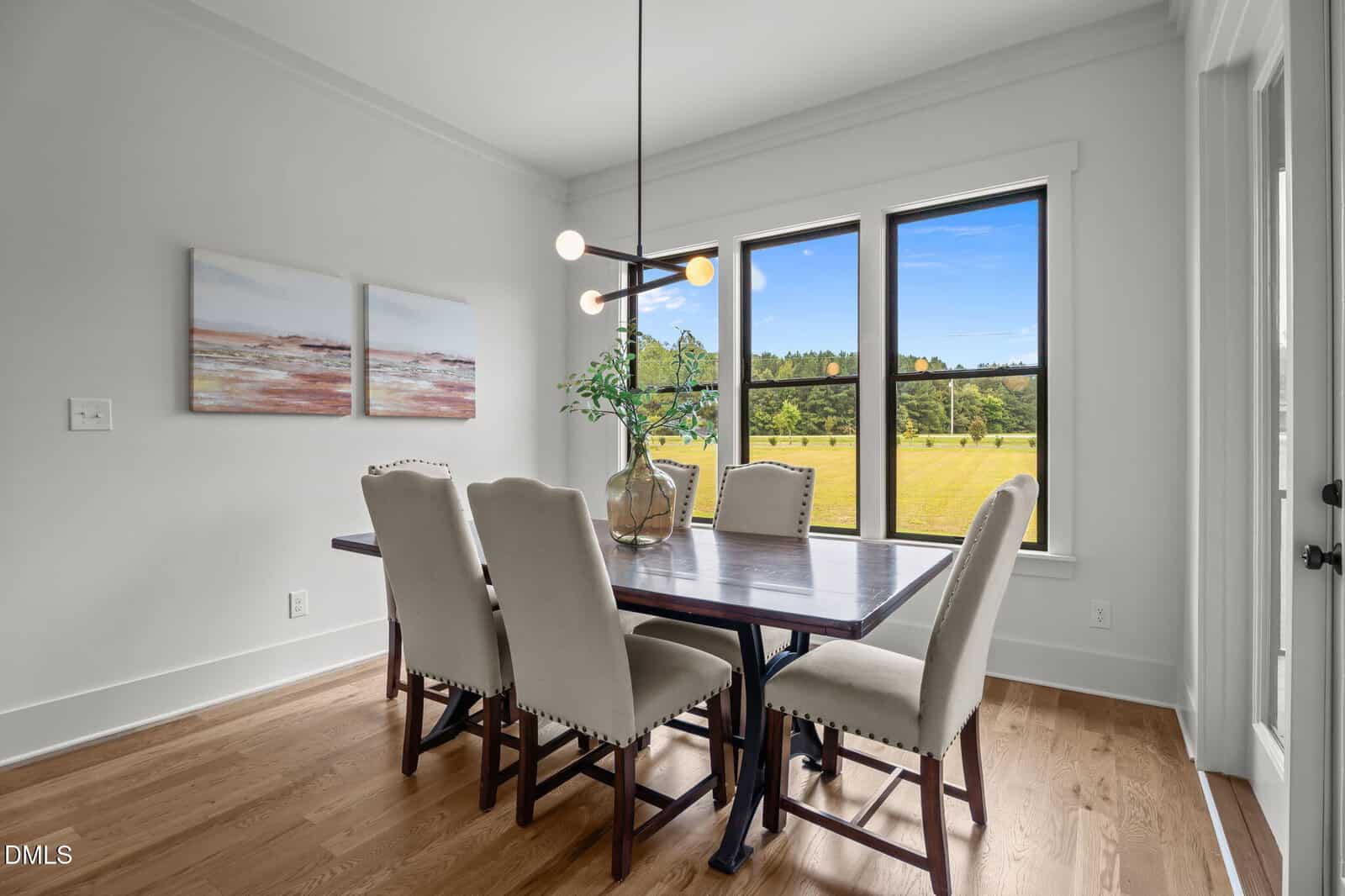 A modern dining room with a wooden table, six upholstered chairs, a glass vase with greenery, abstract wall art, large windows with a scenic outdoor view, and hardwood floors.