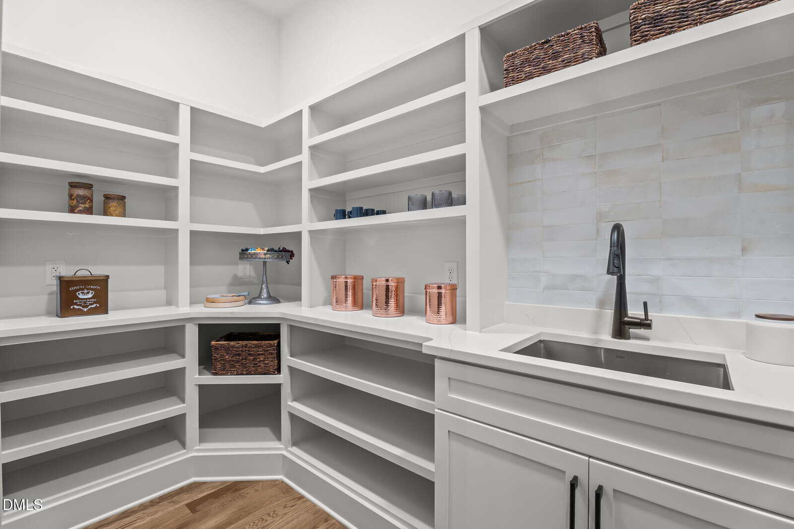 A modern pantry with white shelves holding jars, baskets, copper canisters, candles, and a cake stand. The room features a countertop with a built-in sink and light wood flooring.