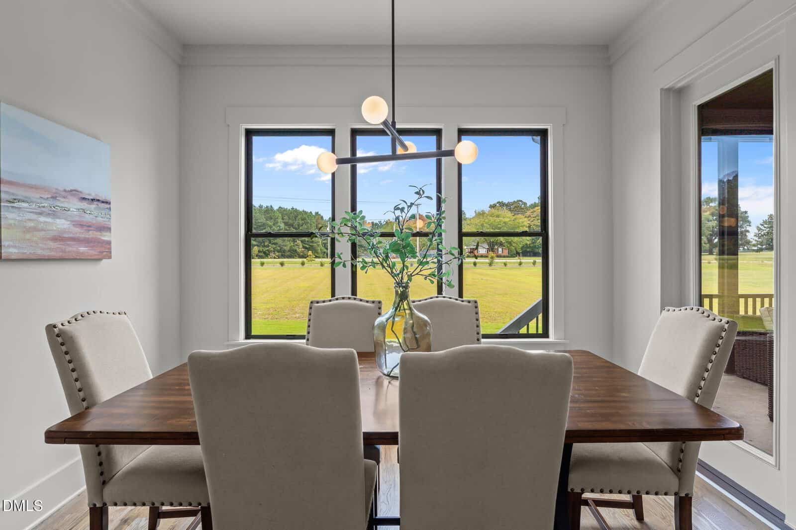 A bright dining room with a wooden table, six beige upholstered chairs, a modern light fixture, and a vase with greenery. Large windows offer a view of a grassy field and trees outside.