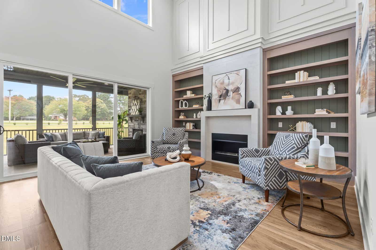 Modern living room with a high ceiling, built-in shelves, abstract art above a fireplace, two patterned chairs, a light gray sofa, a round coffee table, and large sliding doors opening to a patio with a scenic outdoor view.