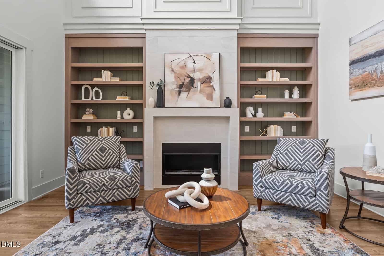 Modern living room with two patterned armchairs, a round coffee table, built-in bookshelves, and a fireplace with abstract art above. Decor includes books, vases, and decorative objects in neutral tones.