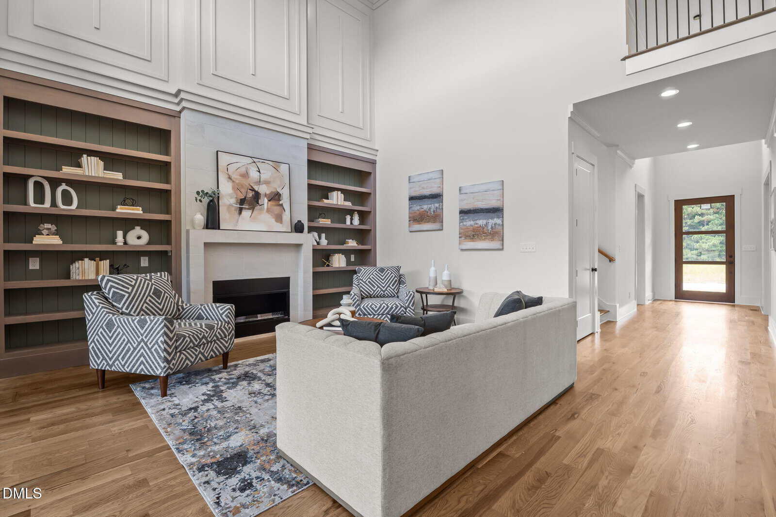 Modern living room with high ceilings, built-in bookshelves, a fireplace, patterned chairs, a light gray sofa, and artwork on the wall. Hardwood floors and hallway leading to a glass door with outdoor view.