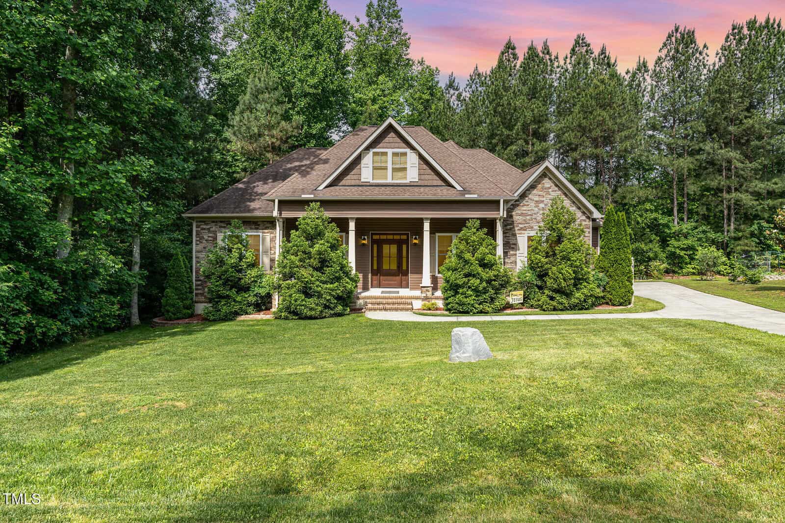 A single-story house with brown siding, stone accents, and a covered front porch is surrounded by green trees and bushes. There is a large lawn and a stone marker in front, with a driveway on the right.