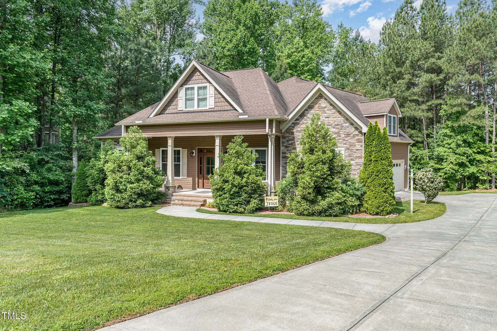 A two-story house with brown siding and stone accents, a covered front porch with columns, surrounded by trees and shrubs, set on a neatly manicured lawn with a curved concrete driveway.