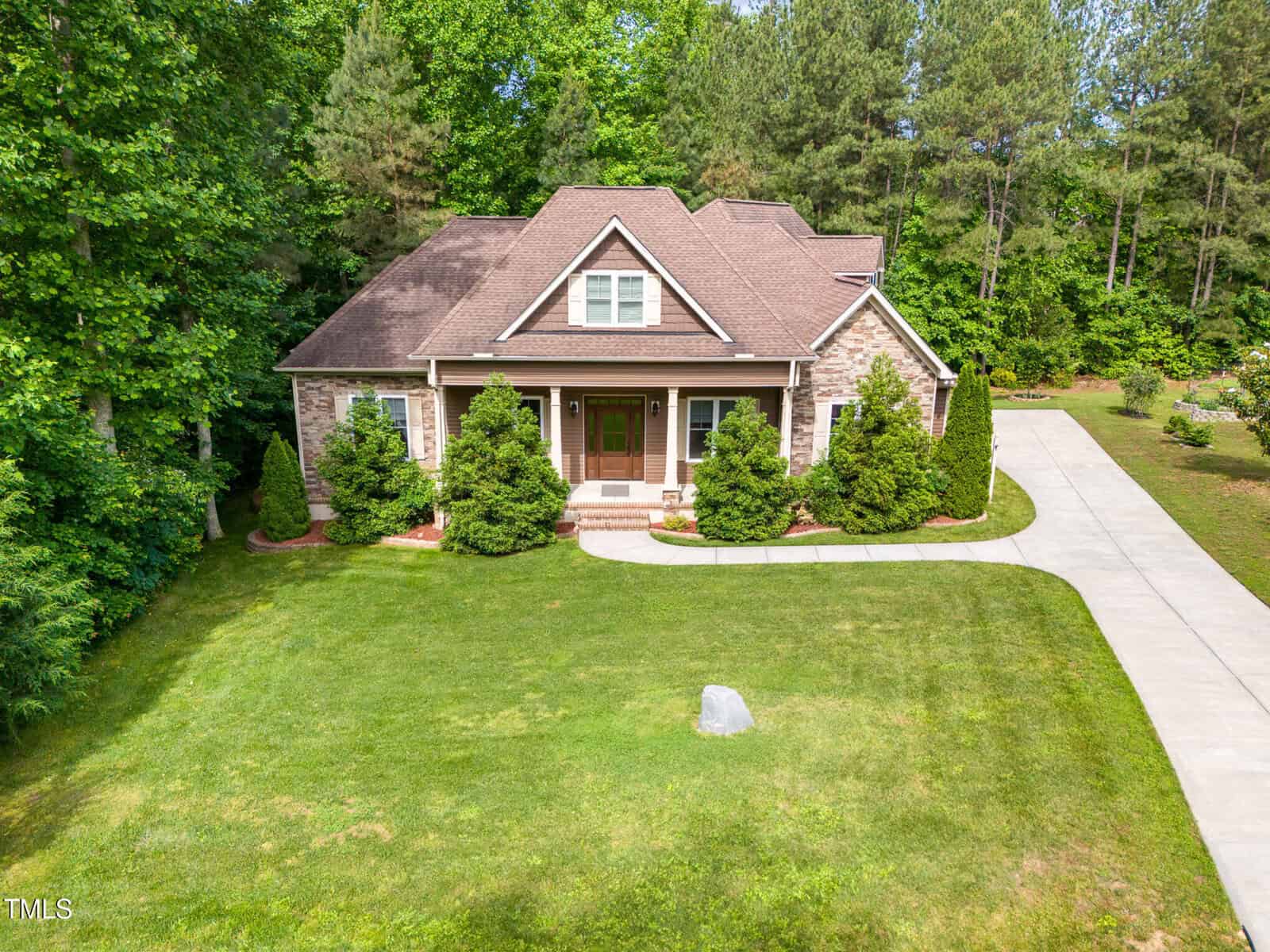 A single-story house with a brown roof and brick exterior sits on a well-kept lawn, surrounded by trees. A concrete driveway runs along the right side of the house, and shrubs line the front porch.