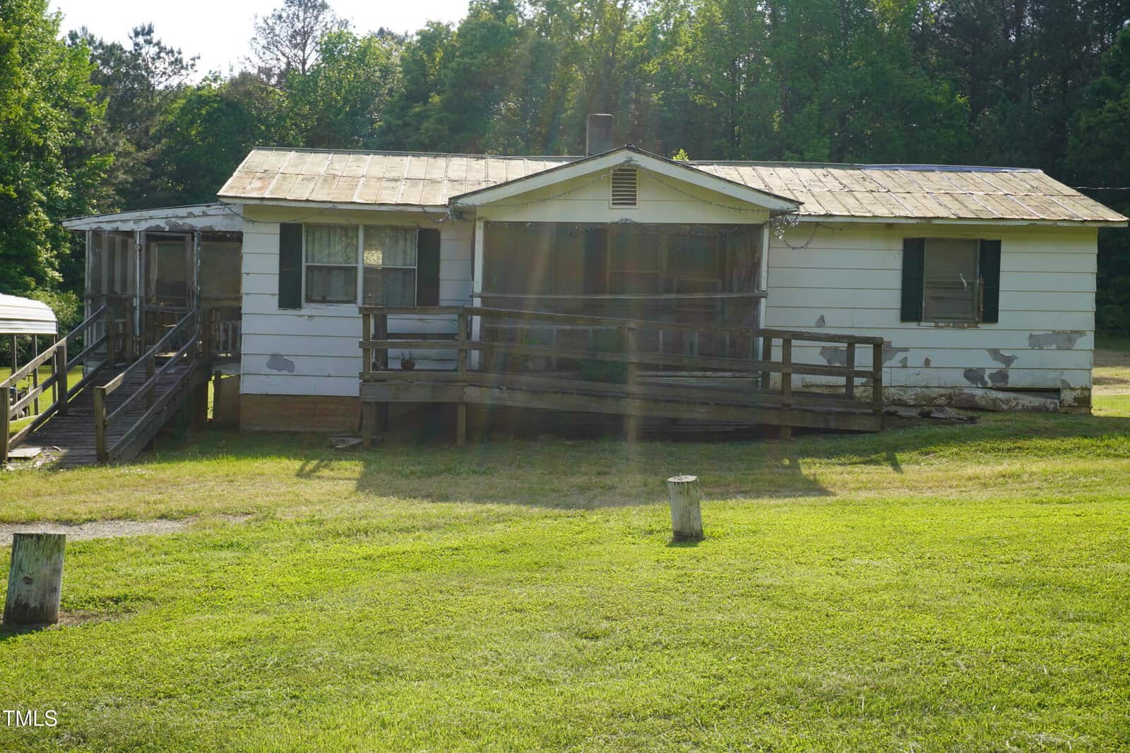 A small, single-story house with a metal roof, wheelchair ramps, peeling paint, and green shutters sits on a grassy lawn along 691 Nc 58 Hwy in Castalia, with trees in the background and sunlight streaming down.
