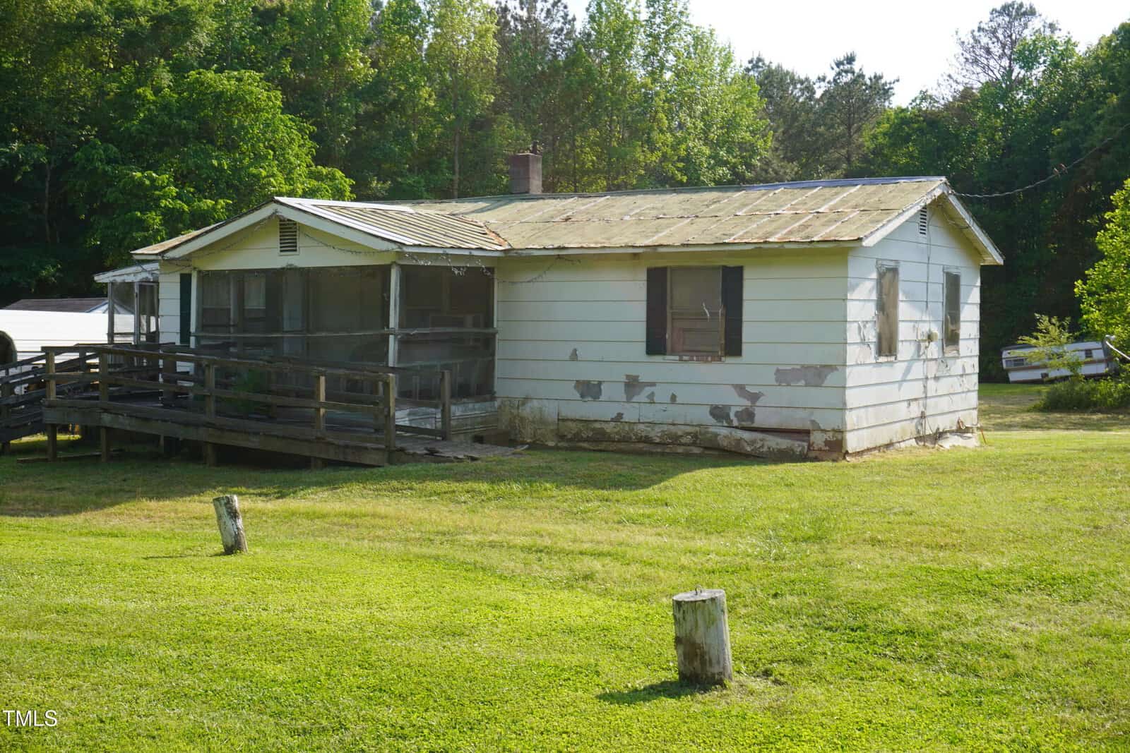 A small, single-story house with peeling white paint, a rusted metal roof, and a wooden ramp. The house is surrounded by grass and trees, with a few posts in the yard.