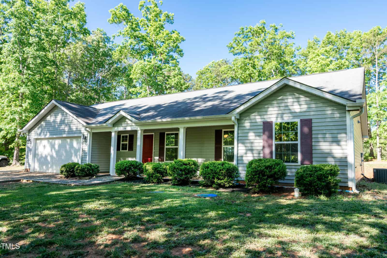 Single-story gray house with a gabled roof, white trim, and dark shutters. There are several windows, a front porch with columns, and neatly trimmed bushes in front. Lush green trees and grass surround the home.