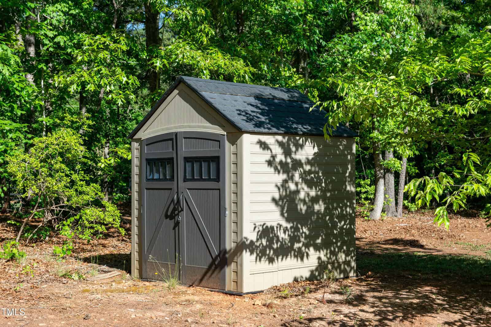 A small beige storage shed with double dark gray doors and a shingled roof sits on bare ground surrounded by green trees and foliage. Sunlight casts shadows on the shed and the ground.