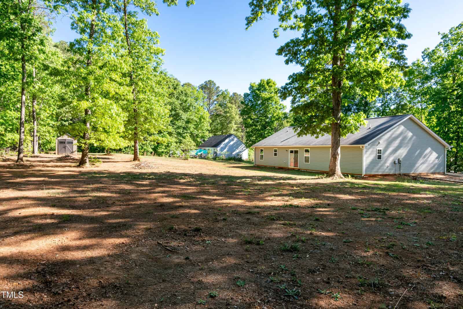 A backyard with sparse grass and large trees, showing a gray single-story house on the right and a small shed in the distance on the left, under a clear blue sky.