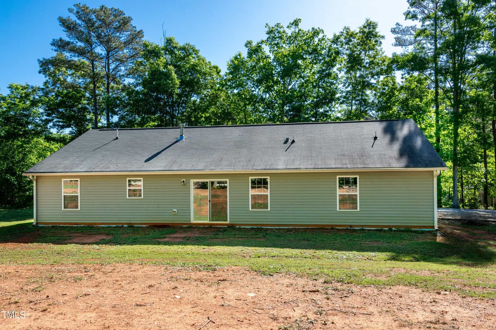 A single-story house with light green siding, six windows, and a glass sliding door. The yard is bare dirt with some grass, and there are trees in the background under a clear blue sky.
