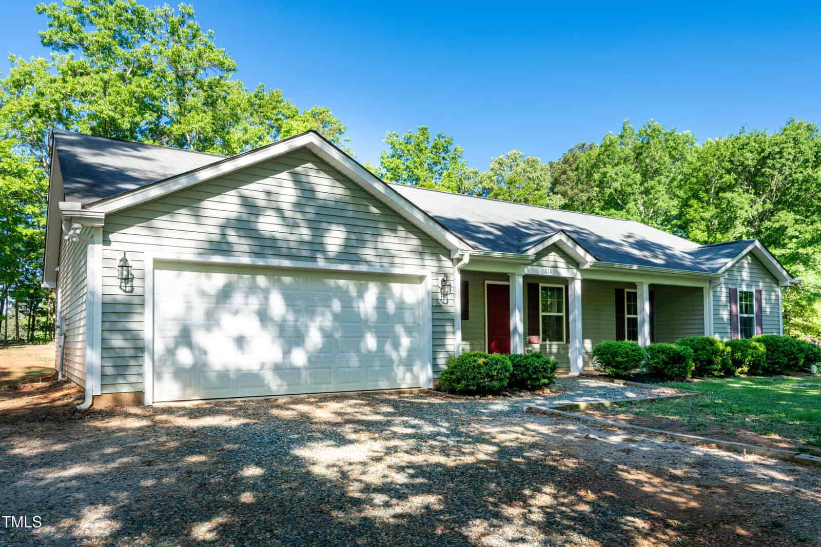 A single-story house with light gray siding, a two-car garage, front porch with columns, and a well-maintained gravel driveway, surrounded by green trees and shrubs under a clear blue sky.